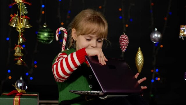 Kid girl in Christmas elf Santa Claus helper costume doing shopping online using laptop isolated on coronavirus quarantine lockdown. Child having video chat browsing on social media. New Year holidays