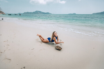 Fototapeta na wymiar Young attractive blonde woman in a blue swimming suit lying on a sandy beach against the sea background. Summer vacation concept