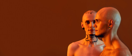 Front and side view of young half naked twin brothers with tattoos and piercings posing together, standing isolated over orange background