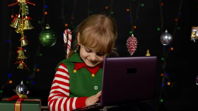 Child looking for presents, gift for friends and family, doing shopping online using laptop isolated on coronavirus quarantine lockdown. Kid girl in Christmas elf Santa Claus helper costume. New Year