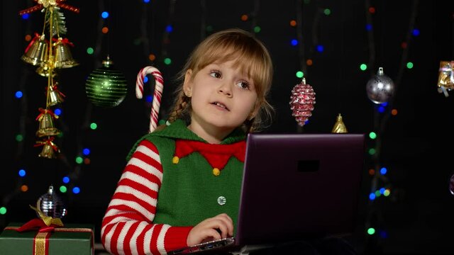 Child looking for presents, gift for friends and family, doing shopping online using laptop isolated on coronavirus quarantine lockdown. Kid girl in Christmas elf Santa Claus helper costume. New Year