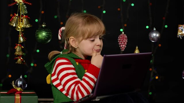 Child looking for presents, gift for friends and family, doing shopping online using laptop isolated on coronavirus quarantine lockdown. Kid girl in Christmas elf Santa Claus helper costume. New Year