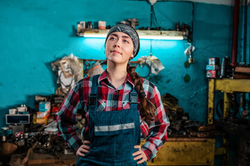 Fototapeta na wymiar Portrait of a young beautiful female mechanic in uniform who poses with her hands on her hips and smiling. Working room in the background. The concept of gender equality