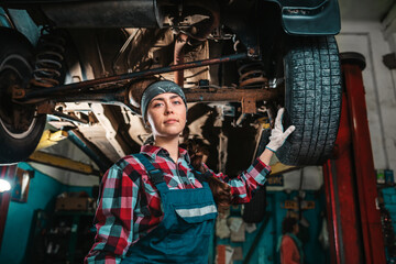 Fototapeta na wymiar Portrait of a young female mechanic in uniform and gloves, posing near a car that is under repair. Bottom view. In the background is an auto repair shop