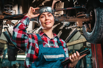 Portrait of a young pretty smiling female mechanic, in a uniform, holding glasses, with a tablet in her hands, poses standing under a car on a lift. Indoors