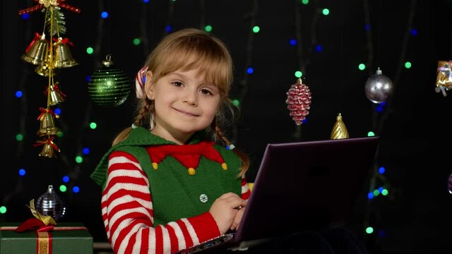 Child looking for presents, gift for friends and family, doing shopping online using laptop isolated on coronavirus quarantine lockdown. Kid girl in Christmas elf Santa Claus helper costume. New Year