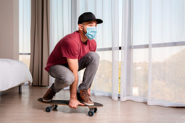 Fototapeta na wymiar A young man in a medical mask poses, sitting on a longboard. Indoor. Home. The concept of quarantine and activity at home