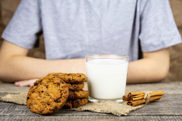 The child sits at a wooden table and holds a glass of milk, oatmeal cookies on the table.