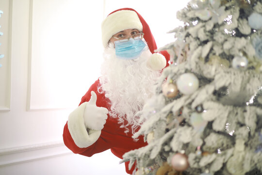 Real Santa Claus and christmas tree on a background, wearing a protective mask, glasses and hat. Christmas with social distance. Covid-19