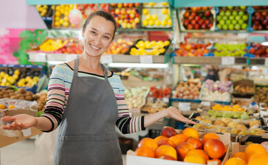 Cheerful woman store worker in apron at fruit department of supermarket