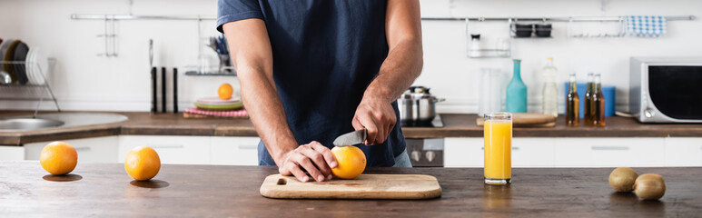 Cropped of man cutting fresh orange near kiwi and glass of orange juice, banner