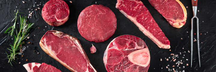 Various cuts of meat panorama, shot from the top on a black background with salt, pepper, rosemary and a carving fork