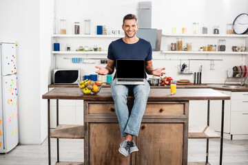  man pointing with hands near laptop with blank screen, orange juice and ripe fruits in kitchen