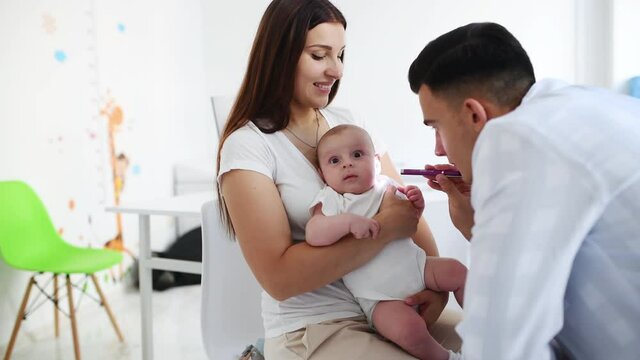 Woman with her son is in the clinic with young doctor that using lighting pen to check ears of baby.
