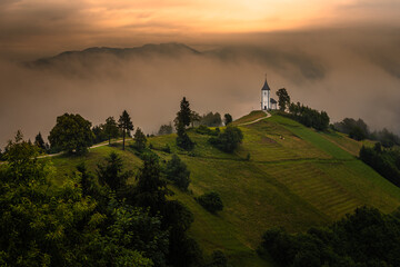 Jamnik, Slovenia - Magical foggy summer morning at Jamnik St.Primoz hilltop church. at sunrise The fog gently goes behind the small chapel with golden sky and Julian Alps at background - obrazy, fototapety, plakaty