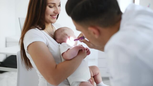 Woman with her son is in the clinic with young doctor that using lighting pen to check ears of baby.