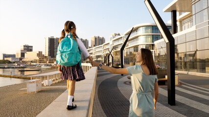 School girl in uniform is walking on railing of seafront while holding hand her young mother, rear view.