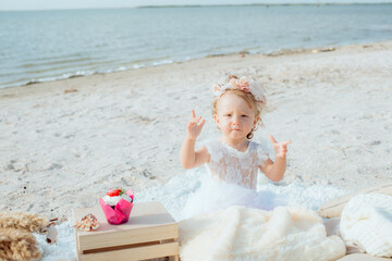 A little cute girl with red curly hair in summer, on the seashore in an airy lace bodysuit and a floral headband devoried with marine details, shells and cotton with birthday cupcake . 1st BIRTHDAY. 
