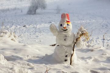 A little cute snowman with a red bucket on his head greets the audience. Winter symbol on the background of a snowy field