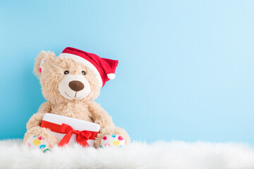 Smiling brown teddy bear with red hat sitting on fluffy fur blanket and holding white Christmas gift box with ribbon. Empty place for text, quote or sayings on light blue wall. Pastel color. Closeup.
