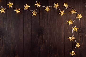 Bright festive background with golden stars glowing of electric lights on elegant dark brown board, frame, top view.
