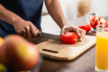 Cropped of man cutting paprika near glass of orange juice on blurred foreground in kitchen