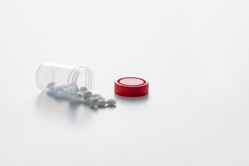 Open transparent pill bottle with some pills on the outside and red cap on the white background