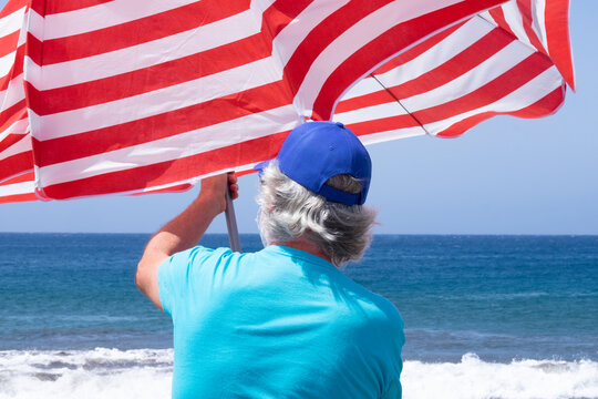 Back View Of Senior Man With White Hair At The Beach Catching Umbrella Flying Away Because Of The Wind - Blue Horizon Over Water In Summer Vacation - One Active Retired People