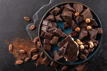 Pieces of dark chocolate, chopped on a round vintage plate, hazelnuts and cocoa beans on a dark...