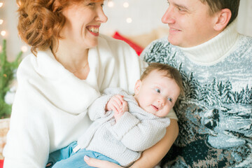 A young family in warm sweaters with a baby daughter in a Christmas bedroom interior decorated with red blankets, pillows, garlands and green pine needles. Family and Children. Christmas mood
