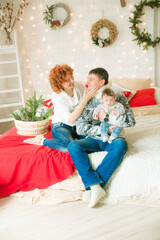 A young family in warm sweaters with a baby daughter in a Christmas bedroom interior decorated with red blankets, pillows, garlands and green pine needles. Family and Children. Christmas mood 