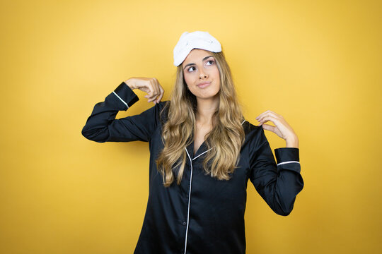 Young pretty woman wearing sleep mask and pajamas over isolated yellow background holding her t-shirt with a successful expression and cockiness