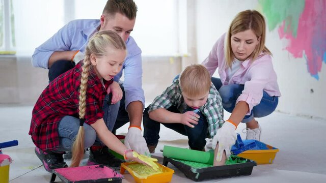 creative family on construction site, girl and boy with down syndrome puts green and yellow acrylic paint on hands from tray with the help of their parents