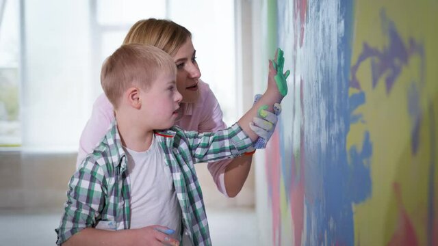 family leisure together at new home, boy with down syndrome with paint on his palm puts a print on a colored wall with the help of mom