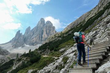 Fototapeta na wymiar Woman with big backpack and sticks, walking up a wooden stairs in high Italian Dolomites. There are many sharp peaks in front of her. She is going up. There are a few trees around. Sunny day. Outdoor