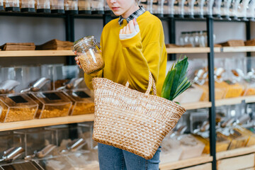 Minimalist vegan style girl with wicker bag and reusable glass coffee cup on background of interior of zero waste shop. Woman doing shopping without plastic packaging in plastic free grocery store.
