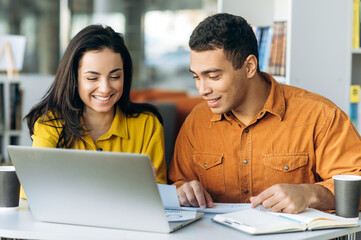 Two colleagues or students, caucasian girl and hispanic guy, are using a laptop while sitting at...