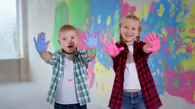 artistic children, happy little girl and cute boy with down syndrome showing to camera colourful painted palms on background of colored wall