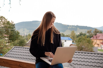 Closeup image of a woman working and typing on laptop while sitting on wooden balcony with green mountains on foggy day with blue sky background