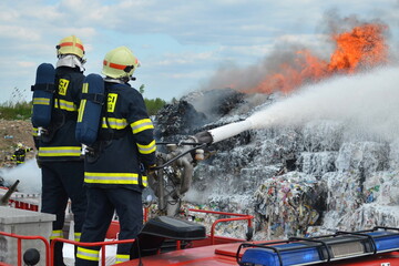 A group of firefighters extinguishes huge landfill fire with flames and black smoke in the background