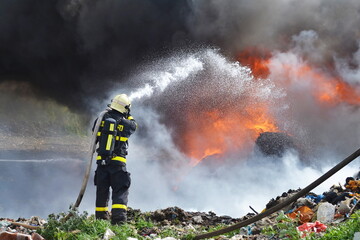 A fireman extinguishes huge landfill fire with flames and black smoke in the background