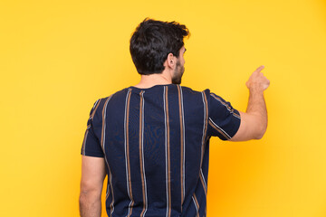 Young handsome man with beard over isolated yellow background pointing back with the index finger