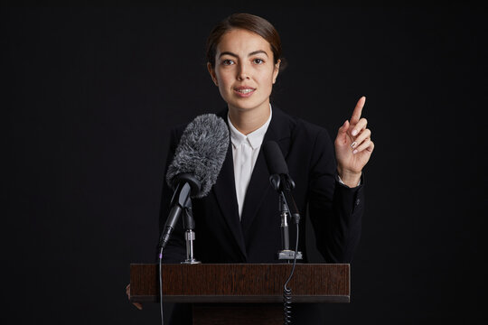 Waist up portrait of young female speaker standing at podium and giving speech to microphone against black background, copy space