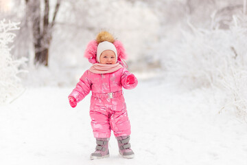 one-year-old sweet baby in pink jumpsuit and boots walking on the white snow in winter. Beautiful children in winter