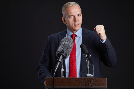 Waist up portrait of mature man standing at podium and gesturing while giving speech to microphone against black background, copy space
