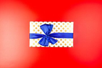 A gift wrapped in white paper with gold circles wrapped in a blue ribbon tied in a bow, isolated on a red background, top view.