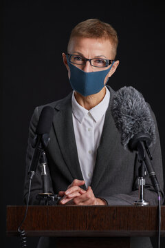 Vertical Portrait Of Mature Woman Wearing Mask While Giving Speech Standing At Podium Against Black Background