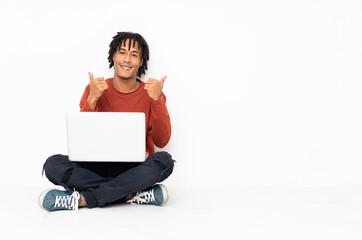 Young african american man sitting on the floor and working with his laptop with thumbs up gesture...