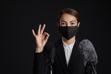 Portrait of young woman wearing mask and showing OK sign while giving speech standing at podium against black background, copy space