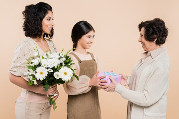 Smiling hispanic girl giving present to grandmother near mother with flowers isolated on beige, three generations of women